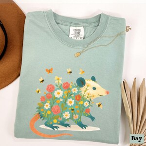 Op de afbeelding: Lichtblauwgroen Comfort Colors T-shirt met een opossum-ontwerp met een bloemenlichaam, omringd door bijen en vlinders. De opossum heeft een gele kop en een lange oranje staart. Een gouden ketting ligt over het shirt.