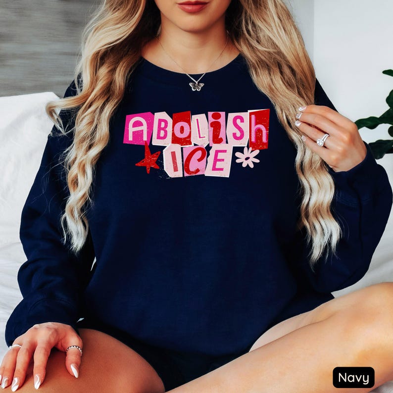 May include: Navy blue sweatshirt with the words "Abolish ICE" in a cut-out style, with pink and red letters. A red star and a white flower are next to the text. The sweatshirt is a solid navy color.