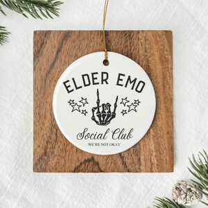 May include: A white ceramic ornament with the text "ELDER EMO Social Club WE'RE NOT OKAY" in black. The ornament features a skeleton hand giving the middle finger and star graphics. It hangs from a gold string and is set on a wooden square.