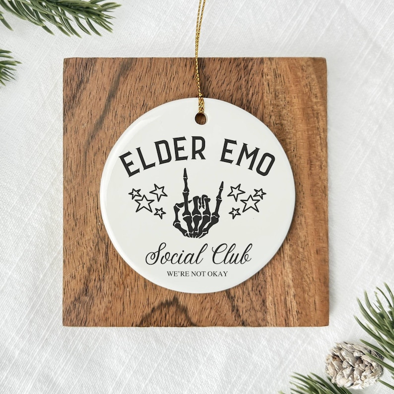 May include: A white ceramic ornament with the text "ELDER EMO Social Club WE'RE NOT OKAY" in black. The ornament features a skeleton hand giving the middle finger and star graphics. It hangs from a gold string and is set on a wooden square.