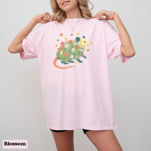 Op de afbeelding: Lichtroze T-shirt met een grillige illustratie van een opossum bedekt met bloemen en omringd door bijen en vlinders. De opossum heeft een lichtgele kop en een lange oranje staart. Het woord "Blossom" staat onderaan.