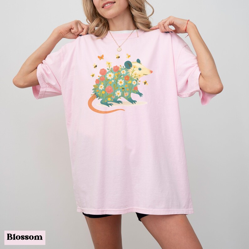 Op de afbeelding: Lichtroze T-shirt met een grillige illustratie van een opossum bedekt met bloemen en omringd door bijen en vlinders. De opossum heeft een lichtgele kop en een lange oranje staart. Het woord "Blossom" staat onderaan.