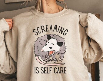 Könnte beinhalten: Ein beiger Pullover mit einem Aufdruck eines Opossums und dem Text "Screaming is Self Care".