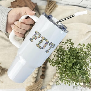 May include: White insulated tumbler with a handle and a lid, featuring the letters "FDT" in a floral pattern. A stainless steel straw with a white tip is inserted into the lid. The tumbler is held by a person.