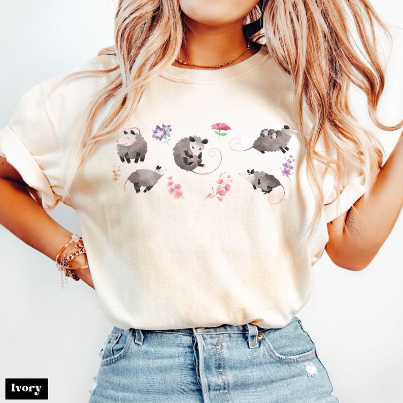 May include: Ivory-colored t-shirt featuring a watercolor design of opossums and flowers. The opossums are depicted in various poses, surrounded by pink, purple, and red floral accents. The shirt has a relaxed fit and is made of a soft material.