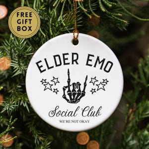 May include: White ceramic ornament with the text "ELDER EMO Social Club WE'RE NOT OKAY" in black. A black skeleton hand with the index and pinky fingers raised is in the center, surrounded by stars. A gold string is attached for hanging.