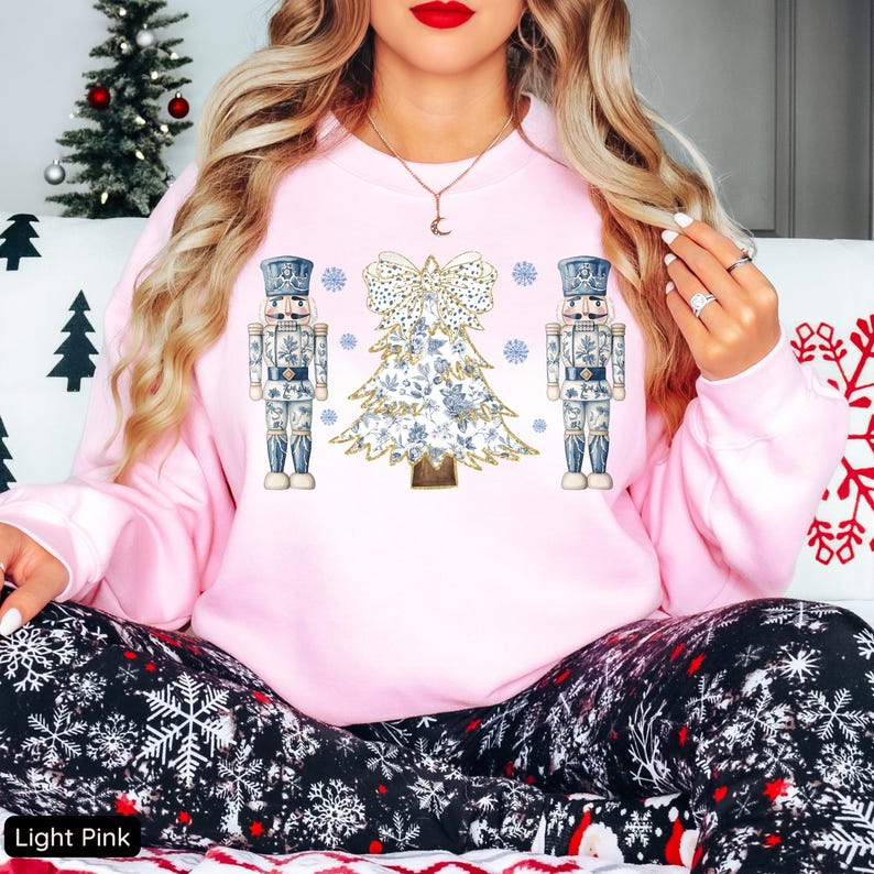 May include: A light pink sweatshirt with a Christmas tree and two nutcracker soldiers printed on the front. The tree is decorated with a white and gold bow and snowflakes.