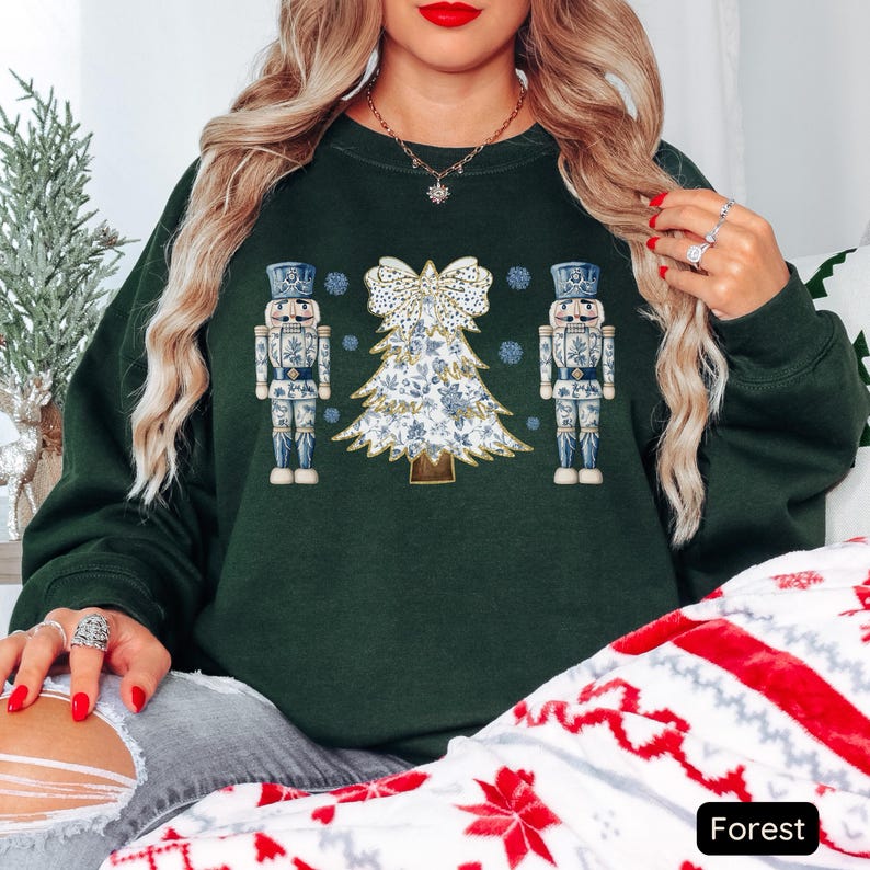 May include: A dark green sweatshirt with a white and blue Christmas tree and two nutcracker figures. The tree is decorated with a white bow and gold accents. The nutcrackers are wearing blue and white uniforms.
