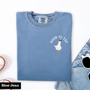 Op de afbeelding: Blauw sweatshirt met ronde hals, met de tekst "BORN TO YAP" en een cartoon gans met zonnebril. Het sweatshirt is gemaakt van zacht materiaal en heeft een "COMFORT COLORS" label.