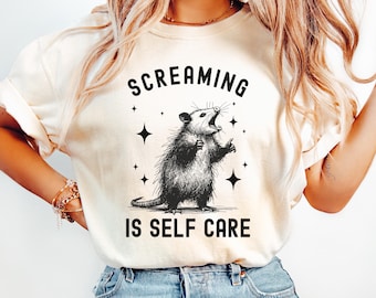 Puede incluir: Una camiseta blanca con una ilustración en blanco y negro de una zarigüeya gritando con estrellas a su alrededor. El texto "Screaming is Self Care" está impreso debajo de la zarigüeya.