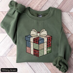 Könnte beinhalten: Militärgrünes Sweatshirt mit einer Grafik einer Patchwork-Geschenkbox und einer cremefarbenen Schleife. Das Geschenkbox-Design enthält Karomuster und unifarbene Muster. Der Text "Military Green" befindet sich unten.
