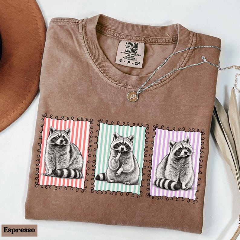 Op de afbeelding: Espresso-kleurig T-shirt met drie wasbeerillustraties in decoratieve kaders. Elke wasbeer wordt in een andere pose afgebeeld tegen een achtergrond van verticale strepen in rood, mintgroen en paars.