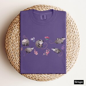 May include: A grape-colored t-shirt featuring a whimsical design of opossums and flowers. The opossums are depicted in various poses, with some carrying babies. The shirt is folded on a woven surface.