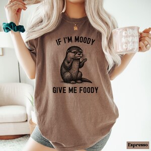 May include: A brown t-shirt with a graphic of an otter and the text "If I'm Moody Give Me Foody".