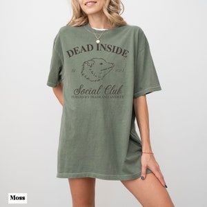 May include: A green oversized t-shirt with a graphic print of an opossum and the text "Dead Inside Social Club Fueled by Trash and Anxiety".