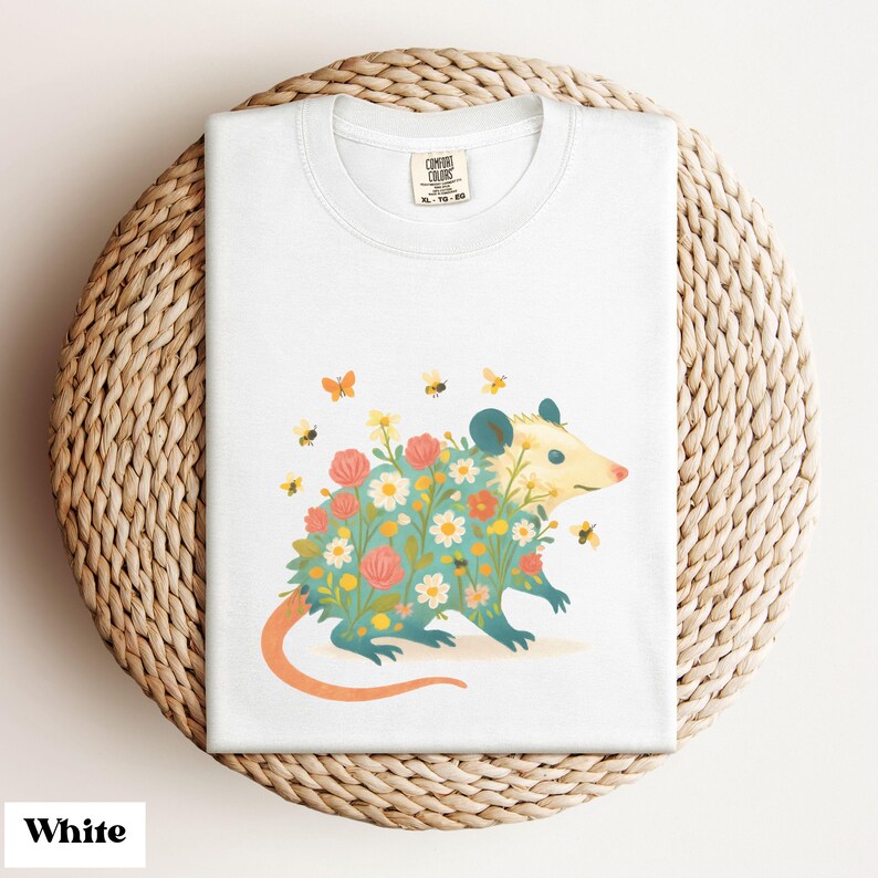 Op de afbeelding: Wit T-shirt met een kleurrijke illustratie van een opossum bedekt met bloemen en omringd door bijen en vlinders. De opossum heeft een lichtblauw lichaam, een roze neus en een oranje staart. Het shirt is op een geweven oppervlak gevouwen.