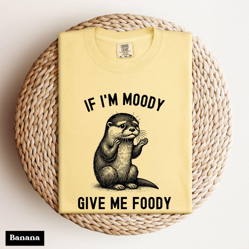 May include: A yellow t-shirt with the text "If I'm Moody Give Me Foody" and a black and white illustration of an otter.
