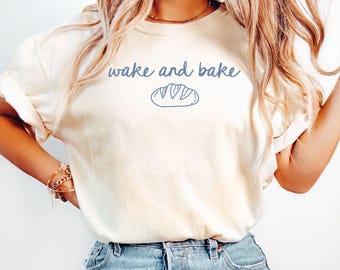 Könnte beinhalten: Elfenbeinfarbenes T-Shirt mit dem Text "wake and bake" in blauer Schrift, begleitet von einer blauen Brotlaib-Illustration. Das Shirt besteht aus einem weichen Material und wird mit blauen Jeans getragen.