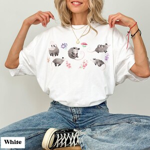 May include: White t-shirt featuring a watercolor design of opossums and floral accents. The opossums are depicted in shades of gray, with pink and purple flowers. The word "White" is printed at the bottom left.