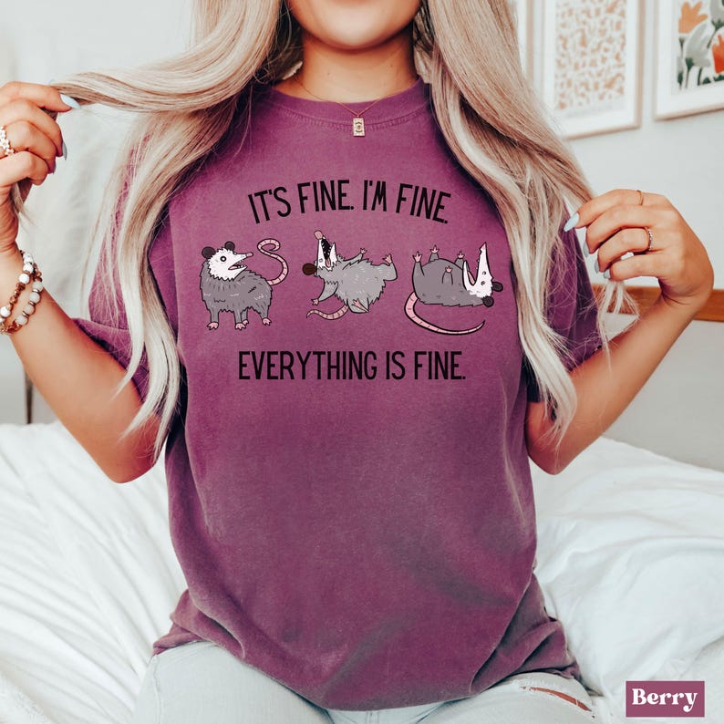 Puede incluir: Una camiseta morada con un gr&aacute;fico de tres zarig&uuml;eyas y el texto "Est&aacute; bien. Estoy bien. Todo est&aacute; bien."