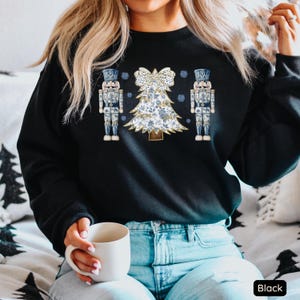 May include: A black sweatshirt with a white and blue Christmas tree design and two nutcracker figures on either side. The tree is decorated with a bow and snowflakes. The sweatshirt is worn by a person with long blonde hair and blue jeans.