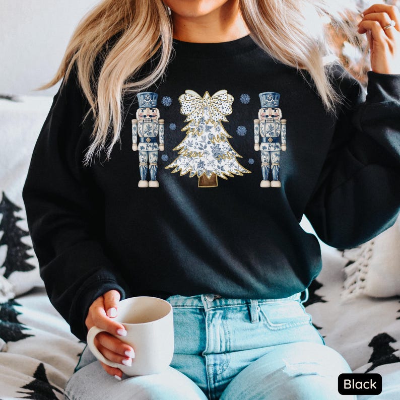 May include: A black sweatshirt with a white and blue Christmas tree design and two nutcracker figures on either side. The tree is decorated with a bow and snowflakes. The sweatshirt is worn by a person with long blonde hair and blue jeans.