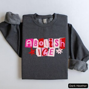 May include: Dark heather gray sweatshirt with the text "Abolish ICE" in a cut-out style, featuring pink and red letters, a red star, and a white flower graphic. The sweatshirt has a crew neck and cuffed sleeves.