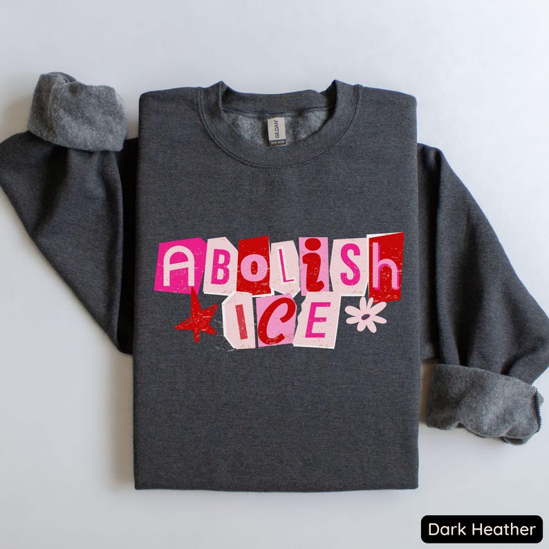 May include: Dark heather gray sweatshirt with the text "Abolish ICE" in a cut-out style, featuring pink and red letters, a red star, and a white flower graphic. The sweatshirt has a crew neck and cuffed sleeves.