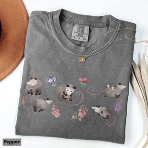May include: A gray Comfort Colors t-shirt featuring a whimsical design of opossums and flowers. The shirt has a crew neck and short sleeves. A silver necklace with a pendant is draped across the shirt. The word "Pepper" is at the bottom left.