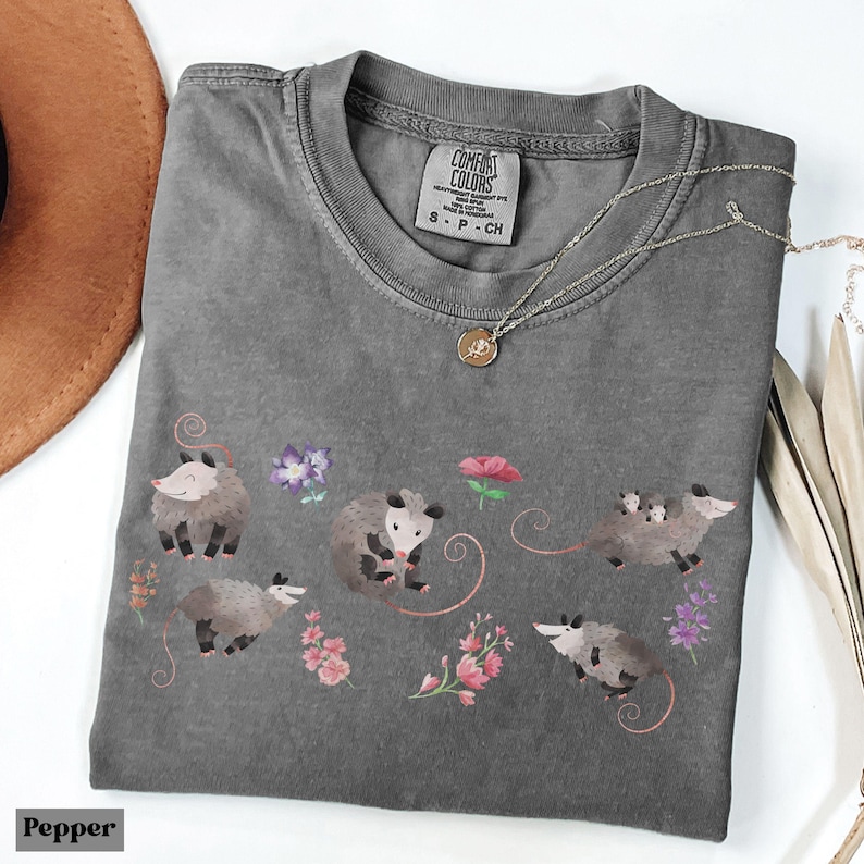 May include: A gray Comfort Colors t-shirt featuring a whimsical design of opossums and flowers. The shirt has a crew neck and short sleeves. A silver necklace with a pendant is draped across the shirt. The word "Pepper" is at the bottom left.