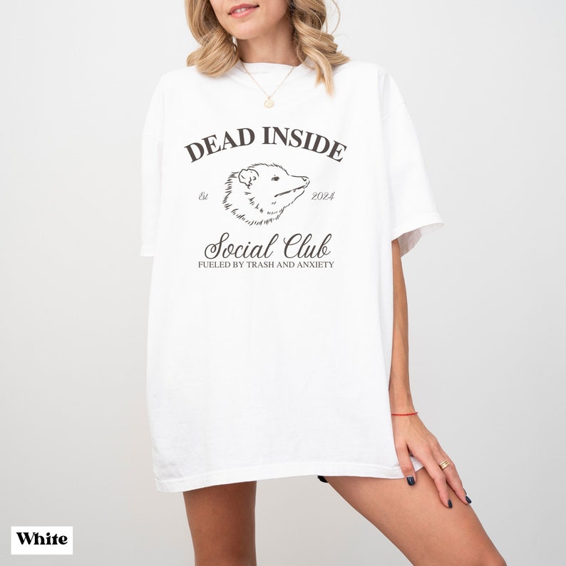 May include: White oversized t-shirt with a black graphic print that says "Dead Inside Social Club est. 2024 Fueled by trash and anxiety".