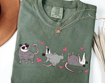 Op de afbeelding: Een groen T-shirt met een afbeelding van drie opossums die een zonnebril dragen en harten vasthouden. Het label staat "COMFORT COLORS".