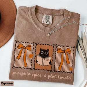 Puede incluir: Camiseta color espresso con un gráfico que presenta un gato negro leyendo un libro, lazos naranjas y el texto "pumpkin spice & plot twists". Un collar plateado con un colgante está sobre la camiseta. La camiseta es talla S.