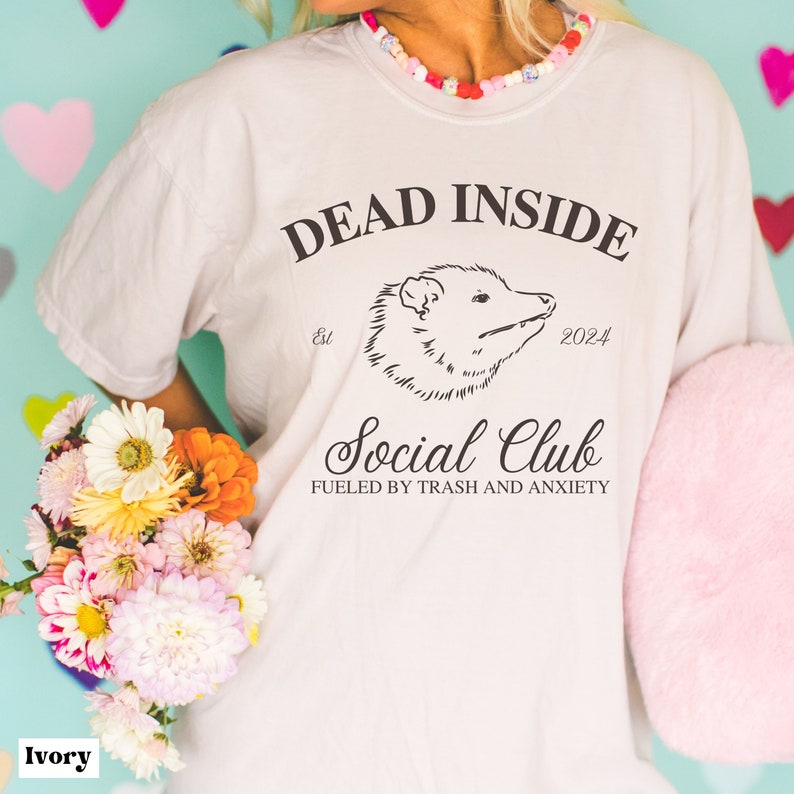 May include: Ivory t-shirt with a black graphic of an opossum and the text "Dead Inside est. 2024 Social Club Fueled by Trash and Anxiety".