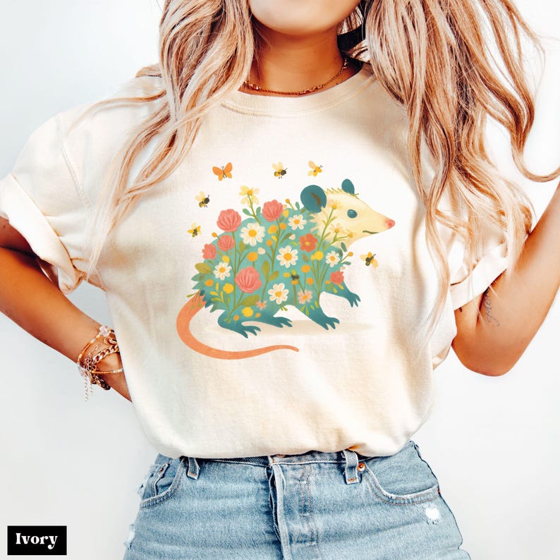 Op de afbeelding: Ivoor kleurig T-shirt met een bloemenbuidelrat ontwerp. De buidelrat is versierd met kleurrijke bloemen en omringd door bijen en vlinders. Het shirt heeft korte mouwen en een ronde hals. Het woord "Ivory" staat onderaan.