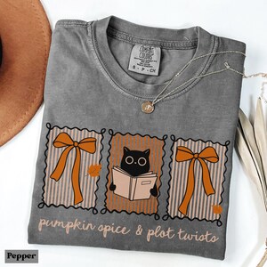 Puede incluir: Una camiseta gris con un gráfico que presenta lazos naranjas, un gato negro leyendo un libro y el texto "pumpkin spice & plot twists". La camiseta tiene cuello redondo y mangas cortas. Un collar plateado con un colgante está sobre la camiseta.