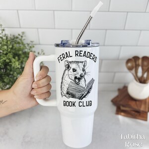 May include: White travel mug with a handle, featuring the text "FERAL READERS BOOK CLUB" and a black and white illustration of a rodent reading a book. Includes a silver straw and a clear lid.
