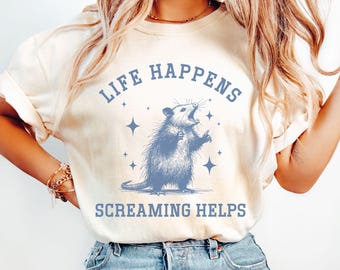 May include: Ivory-colored t-shirt with the text "LIFE HAPPENS SCREAMING HELPS" in blue. The graphic features a blue opossum with its mouth open, surrounded by stars. The shirt is a casual wear item.