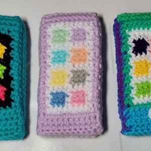 Op de afbeelding: Drie gehaakte telefoonhoesjes met verschillende kleurschema's en patronen. Het eerste hoesje is teal met een zwart-wit rasterpatroon en een kleurrijk vierkant ontwerp in het midden. Het tweede hoesje is lavendel met een wit rasterpatroon en een kleurrijk vierkant ontwerp in het midden. Het derde hoesje is groen met een wit rasterpatroon en een kleurrijk vierkant ontwerp in het midden.