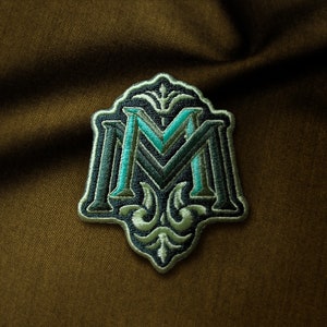 Puede incluir: Un parche bordado verde y azul con la letra "M" en una fuente decorativa. El parche tiene un diseño floral detallado alrededor de la letra.