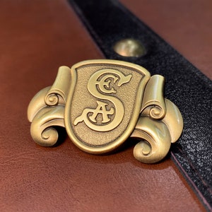 May include: A gold-toned metal pin with a decorative scroll design. The pin features a shield with the letters "S.A." inside.