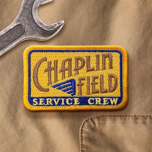 May include: A rectangular patch with the words "CHAPLIN FIELD SERVICE CREW" in brown and blue lettering. The patch has a yellow background and a blue border. A silver spanner is in the upper left corner. The patch is on a tan fabric.
