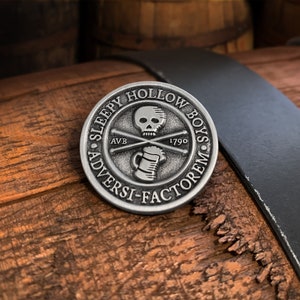 May include: A silver-colored coin with the text "SLEEPY HOLLOW BOYS" encircling a central design. The design includes a skull, crossed bones, a beer mug, and the year 1799. The coin rests on a wooden surface with a black strap.