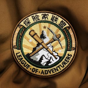 May include: A circular embroidered patch featuring a central mountain graphic with a crossed telescope and pickaxe. The design includes a sunburst background, a dark green border, and the text "LEAGUE OF ADVENTURERS" and Chinese characters.