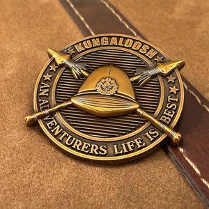 May include: A circular brass-toned metal coin featuring a central pith helmet emblem with two crossed spears. The border displays the text KUNGALOOSH and AN ADVENTURERS LIFE IS BEST with small stars separating the phrases on a lined background.
