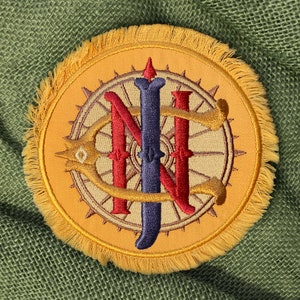 May include: A circular embroidered patch with a gold fringe border. The center features a compass rose design in light tan and brown, overlaid with the letters N, I, and C in red, dark blue, and gold thread, respectively.