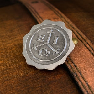 May include: A silver-colored metal seal with the text "E I Co" and a cross design. The seal is round with a scalloped edge and is set against a brown leather background.