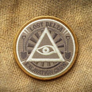 Op de afbeelding: Ronde patch met een bruin en beige ontwerp. De patch heeft een driehoek met een oog, schoppen en de woorden "LOST DELTA EXCAVATION CREW". Het heeft een gouden rand en ligt op een beige stoffen achtergrond.