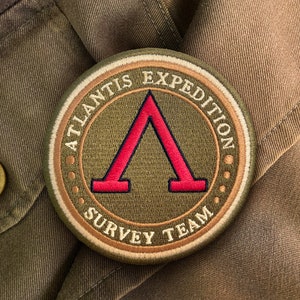 Könnte beinhalten: Runder, gestickter Aufnäher mit dem Text "ATLANTIS EXPEDITION SURVEY TEAM". Er zeigt ein rotes griechisches Lambda-Symbol in der Mitte, umgeben von einem grünen und beigen Rand. Der Aufnäher befindet sich auf einem grünen Stoff.