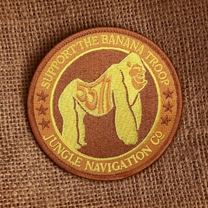 Op de afbeelding: Ronde geborduurde patch met een gele gorilla op een bruine achtergrond. De tekst "SUPPORT THE BANANA TROOP" en "JUNGLE NAVIGATION CO" staan in het geel rond de gorilla. De gorilla heeft het nummer "55" op zijn rug.